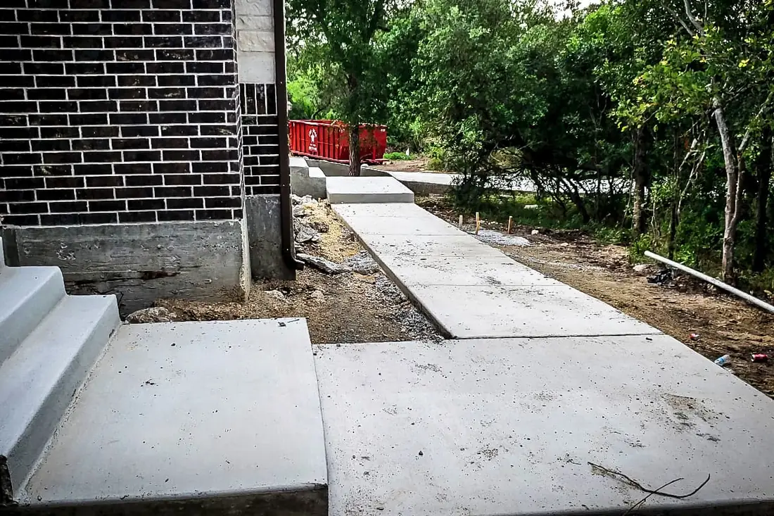 Freshly poured concrete steps beside a new sidewalk at a San Antonio new build, forms just stripped, brick wall behind